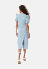 Blue gingham midi dress with puff sleeves, back slit, and fitted waist. Paired with strappy white high-heeled sandals.