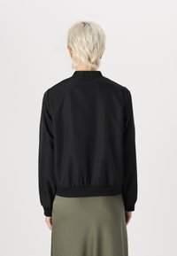 Svart bomberjakke laget av glatt stoff. Har stående krage, ribbestrikkede mansjetter og en tettsittende midje. Minimalistisk design.