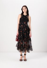 Black floral midi dress with layered ruffles, lace accents, and semi-sheer fabric. Model accessorized with a red handbag and black sandals.