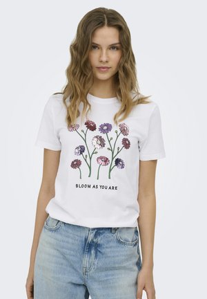 Giovane donna con capelli mossi che indossa una t-shirt bianca con fiori colorati e testo "Bloom as you are,", abbinata a jeans azzurri chiari.