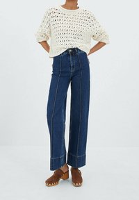 Slonovinový pletený svetr s otevřeným vzorem, doplněný o kalhoty širokého střihu s vysokým pasem z tmavého denimu s kontrastním prošitím a zkrácenými lemy.