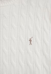 Weißes Strickstoff mit Kabelmuster und einem kleinen bestickten Polospieler in Beige, der auf der unteren rechten Seite einen Schläger hält.