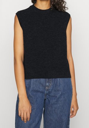 Mouwloze zwarte gebreide top gedragen door persoon met krullend haar, gecombineerd met hoge taille blauwe spijkerbroek tegen een effen achtergrond.