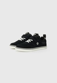 Baskets en suède noir avec des lacets blancs, une sangle en velcro et une semelle en caoutchouc blanc. Présente un logo près du haut et un accent sur le côté.