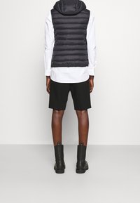 Persona de pie vestida con un chaleco con capucha acolchado negro, una camisa blanca de manga larga, pantalones cortos negros hasta la rodilla y botas negras hasta el tobillo, de espaldas.