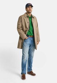 Beige trenchcoat, groene poloshirt, blauwe jeans, bruine laarzen. De jas heeft knoopsluitingen en een kraag; de hoed is marineblauw met rode letters.