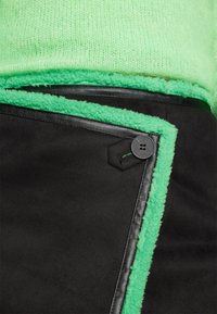 Gros plan d'une jupe noire avec bordure en polaire verte, détail de bouton et une ceinture de pull tricoté vert clair.