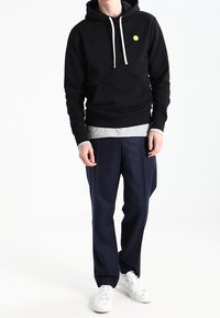 Svart hoodie med en gul logotyp, grå långärmad tröja under, marinblå randiga byxor och vita sneakers.