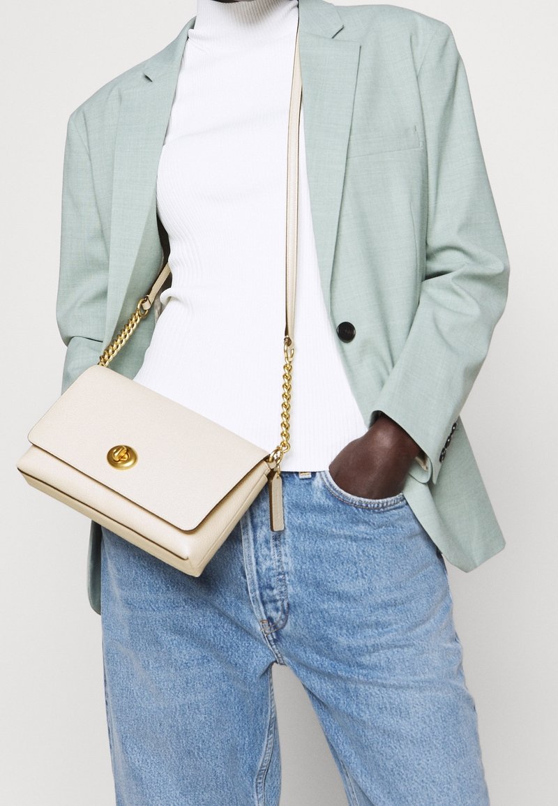 Persona che indossa un blazer verde chiaro, un dolcevita bianco a coste, jeans blu e porta una borsa a tracolla beige con catena dorata e chiusura.