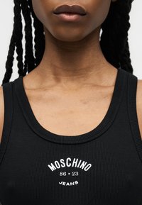 Κοντινό ενός ατόμου που φοράει μαύρο ριπ αμάνικο με λευκό κείμενο "MOSCHINO 86•23 JEANS" στο στήθος.