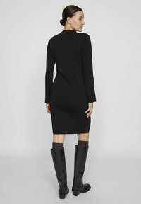 Črna obleka z dolgimi rokavi, prilegajočega kroja, do kolen. Usklajena z visokimi, teksturiranimi črnimi škornji in sivimi nogavicami do kolen.