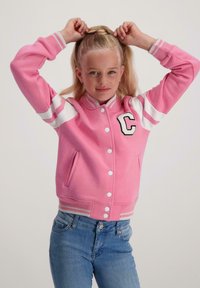 Jong meisje met blond haar die een paardenstaart vastbindt, gekleed in een roze varsityjack met witte strepen en de letter "C" en blauwe jeans, tegen een witte achtergrond.