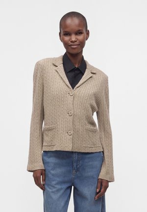 Person iført en beige kabelstrikket cardigan med knapper over en sort skjorte og blå jeans, stående mod en ensfarvet hvid baggrund.