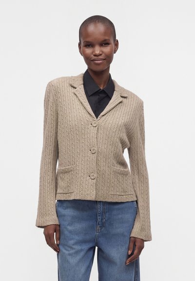 Personne portant un cardigan beige en maille torsadée avec des boutons, sur une chemise noire et un jean bleu, debout devant un fond blanc uni.