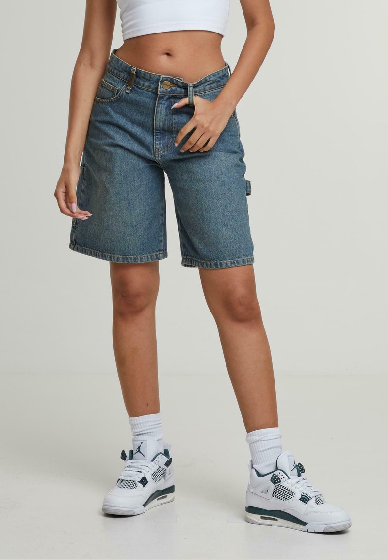 Gaiši zili džinsa īsās bikses ar brīvu piegriezumu, kurām ir piecas kabatas un klasiskā rāvējslēdzēja un pogas aizdare. Valkātas ar baltām sporta apaviem.