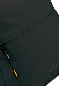 Mochila verde oscuro con tejido texturizado, un bolsillo con cremallera y un pequeño acento amarillo. Incluye una etiqueta de marca con el logo "Bench".