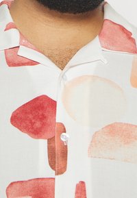 Camicetta a maniche corte in tessuto leggero con sfondo crema e forme astratte rosse e pesca. Collo classico e dettaglio con chiusura a bottoni.