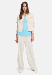 Light beige denim jacket with two front pockets, paired with a blue v-neck top and cream wide-leg pants, complemented by white sneakers.