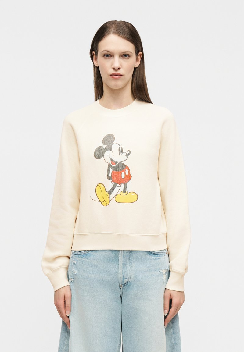 Γυναίκα με κρεμ φούτερ με γραφικό vintage στυλ του Mickey Mouse και ανοιχτό μπλε τζιν με ψηλή μέση, μπροστά σε λευκό φόντο.