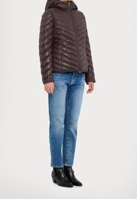 Veste rembourrée matelassée brune avec capuche, présentant un motif en chevrons, associée à un jean bleu clair et des bottes à talons noires.