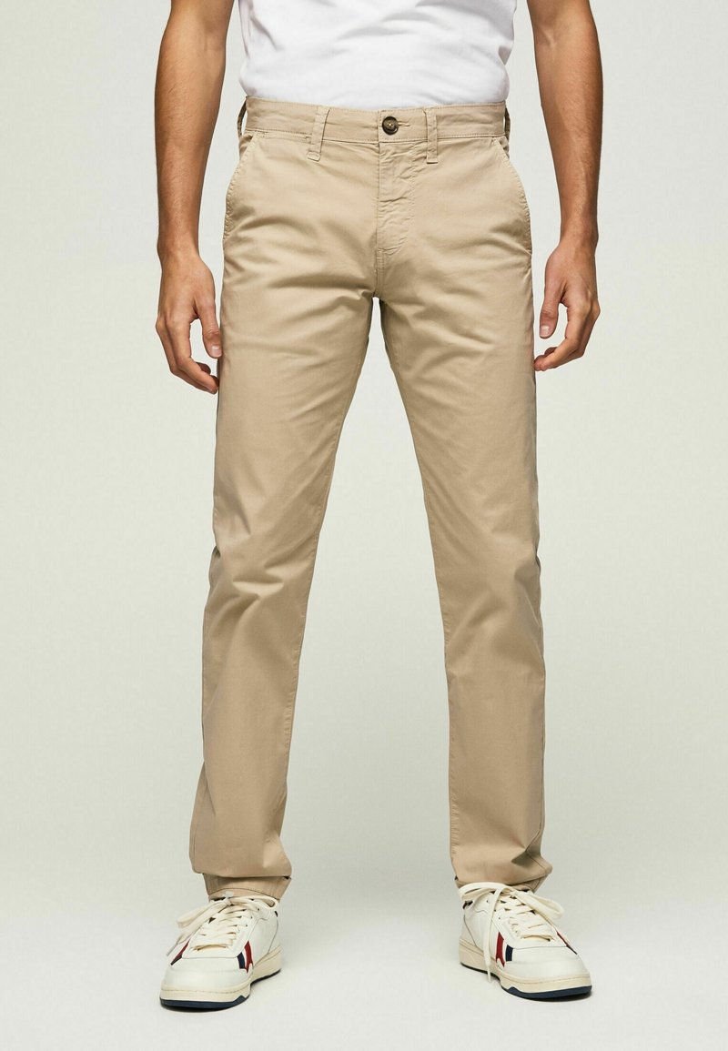 Homme portant un pantalon beige droit, des baskets blanches avec des rayures bleues et rouges, et une chemise blanche, se tenant devant un fond uni.