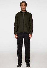 Olivgrön zip-up-jacka med platt krage och framficka, kombinerad med svarta byxor och mörka skor. Tyget verkar ha struktur.