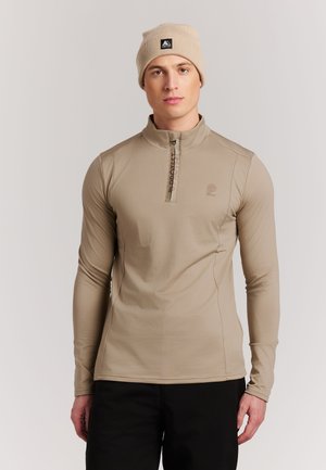 Man in een beige top met lange mouwen en rits en bijpassende muts, staand tegen een eenvoudige lichte achtergrond.