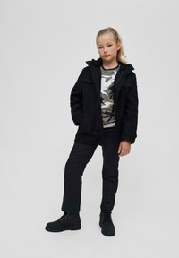 Črna jakna s kapuco obrobljeno s krznom, pod njo kamuflaža srajca, črne cargo hlače in črne čevlje z vezalkami. Model stoji ob enobarvni ozadju.