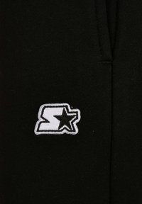 Juodas audinys su minkšta tekstūra, kuriame išsiuvinėtas baltas logotipas, sudarytas iš „S“ ir žvaigždutės, esantis šone.