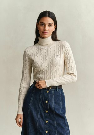Vrouw met lang donker haar draagt een crèmekleurige kabelgebreide coltrui en een blauw denim rokje met hoge taille, gouden knopen en een zwarte riem.