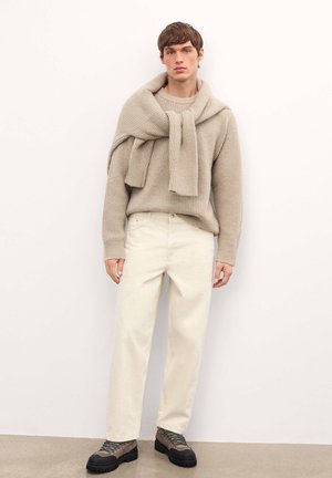 Jeune homme portant un pull en tricot beige avec un pull assorti drapé sur les épaules, un pantalon blanc cassé et des bottes de randonnée sombres, se tenant contre un mur blanc.
