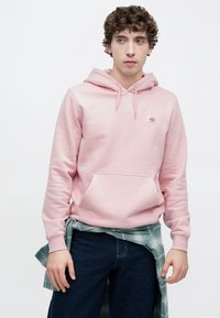 Ανοιχτό ροζ φούτερ με κουκούλα από απαλό ύφασμα, με μπροστινή τσέπη και κορδόνια. Συνδυάζεται με σκουρο μπλε τζιν και πράσινο καρό πουκάμισο δεμένο γύρω από τη μέση.