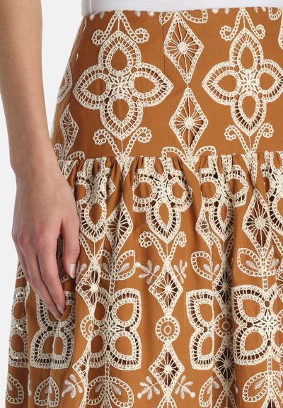 Mână odihnindu-se lângă o fustă maro cu modele florale și geometrice complicate croșetate în culoarea crem.