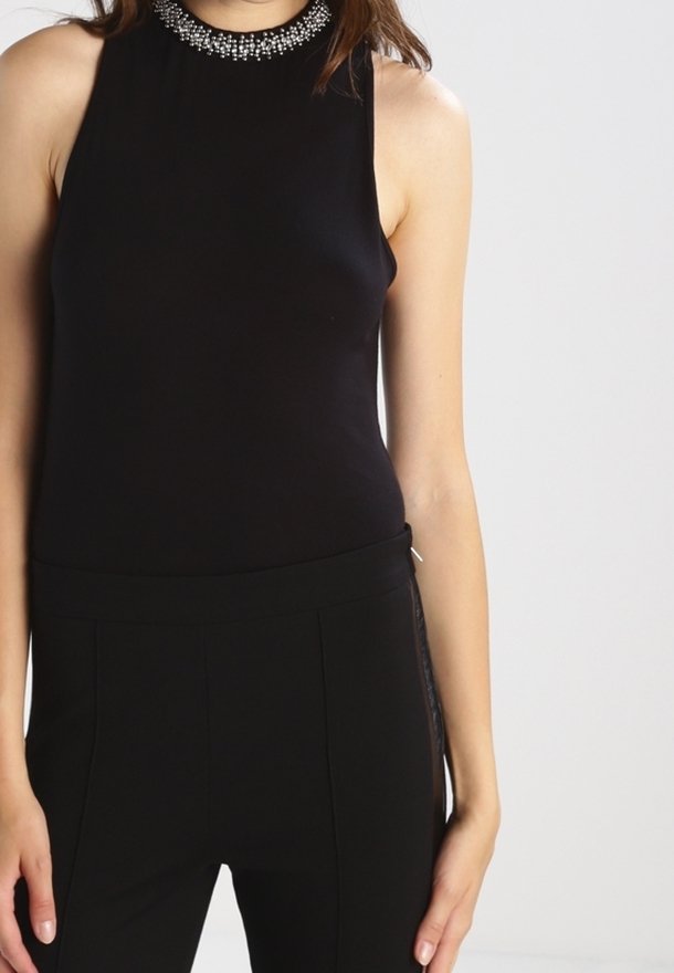 Femme portant un haut noir sans manches avec un col montant orné de bijoux et un pantalon noir ajusté, debout devant un fond clair.