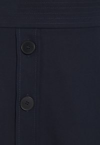 Marinfärgad tyg med strukturerad yta, prydd med två svarta knappar för stängning. Subtil sömdetalj längs den övre kanten.