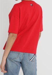 Camiseta de algodón roja con un corte relajado, mangas cortas y escote redondo, con una etiqueta de logo en la parte posterior y jeans de mezclilla azul.
