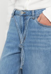 Ljusblå jeans i denim med knytning i midjan, utrustade med klassiska fickor och synliga sömmar. Tyget verkar mjukt med en lätt textur.