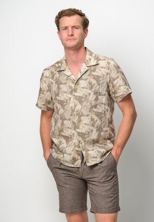 Camisa de manga corta con patrón floral beige, de tela ligera. Combinada con pantalones cortos marrones texturizados. El modelo está de pie con las manos en los bolsillos.