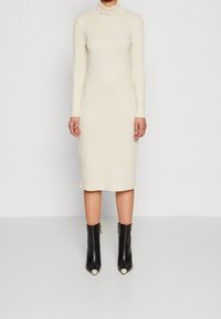 Robe en maille côtelée crème à manches longues avec un col montant, longueur jusqu'aux genoux, associée à des bottines noires avec embouts métalliques.