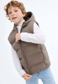 Rjav podloženi brezrokavnik s kapuco, sprednjim zadrgo, nošen preko bele srajce. Ima gladko teksturo in pravokotno prešito strukturo.