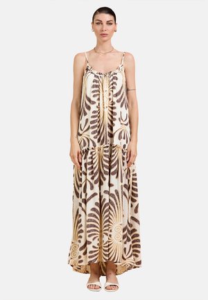 Donna che indossa un vestito lungo beige senza maniche con motivi astratti di foglie marroni e sandali bianchi aperti, in piedi davanti a uno sfondo semplice.