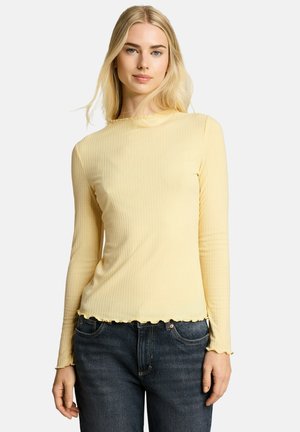 Jonge vrouw met lang blond haar die een lichtgele geribbelde top met lange mouwen en donkerblauwe spijkerbroek draagt tegen een effen achtergrond.
