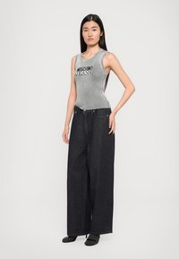 Γκρι ριγέ αμάνικο μπλουζάκι με κείμενο "MOSCHINO JEANS", συνδυασμένο με φαρδιές τζιν παντελόνες σε σκούρο χρώμα. Μοντέλο που φορά μαύρες παντόφλες.