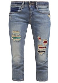Jean en denim bleu clair avec des zones usées révélant un tissu turquoise, rouge et noir en dessous, avec des poches avant et une fermeture par bouton.