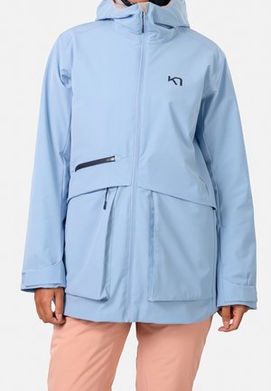 Persoon die een lichtblauwe capuchonjas draagt met zwarte ritssluitingen in de zakken en lichtroze broek, getoond van nek tot heupen.