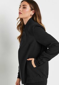 Czarna kurtka bomberka z zamkiem z przodu, elastycznymi mankietami, bocznymi kieszeniami oraz gładką teksturowaną tkaniną.