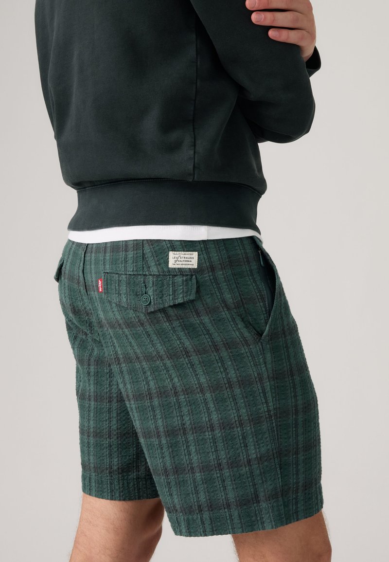 Persona che indossa pantaloncini verdi a quadri con tasca posteriore abbottonata e felpa nera, in piedi con le braccia incrociate su uno sfondo semplice.