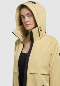 Gul vattentät jacka med dragkedja fram, hög krage och justerbar huva. Har svarta detaljer och en elegant design.