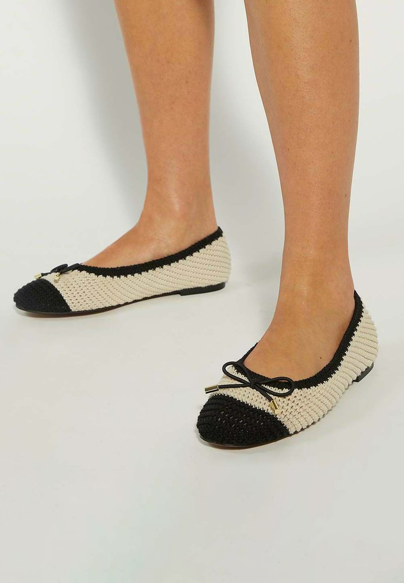 Knit ballet flats featuring a black toe cap and trim with a cream body, adorned with a small black bow and gold accents on the bow.