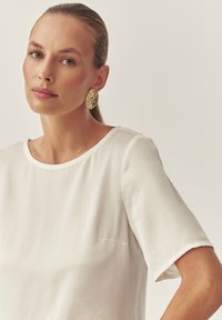 Top blanco de manga corta con un escote redondo, confeccionado en una tela suave y con un ligero brillo. Complementado con pendientes de oro texturizados.